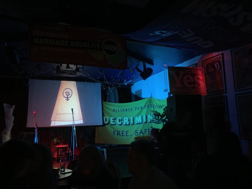 A dark stage with no people on it. Behind the stage are a projected photo of a banner with an icon combining the raised fist of protest and the women's symbol, with a circle above a cross. To the right of the image is a yellow banner whose words are not entirely legible but reads that reads "Alliance for Choice Derry - Decriminalize - Free. Safe. Legal." In the foreground are the backs of several people's heads, waiting for a musician to come onstage.