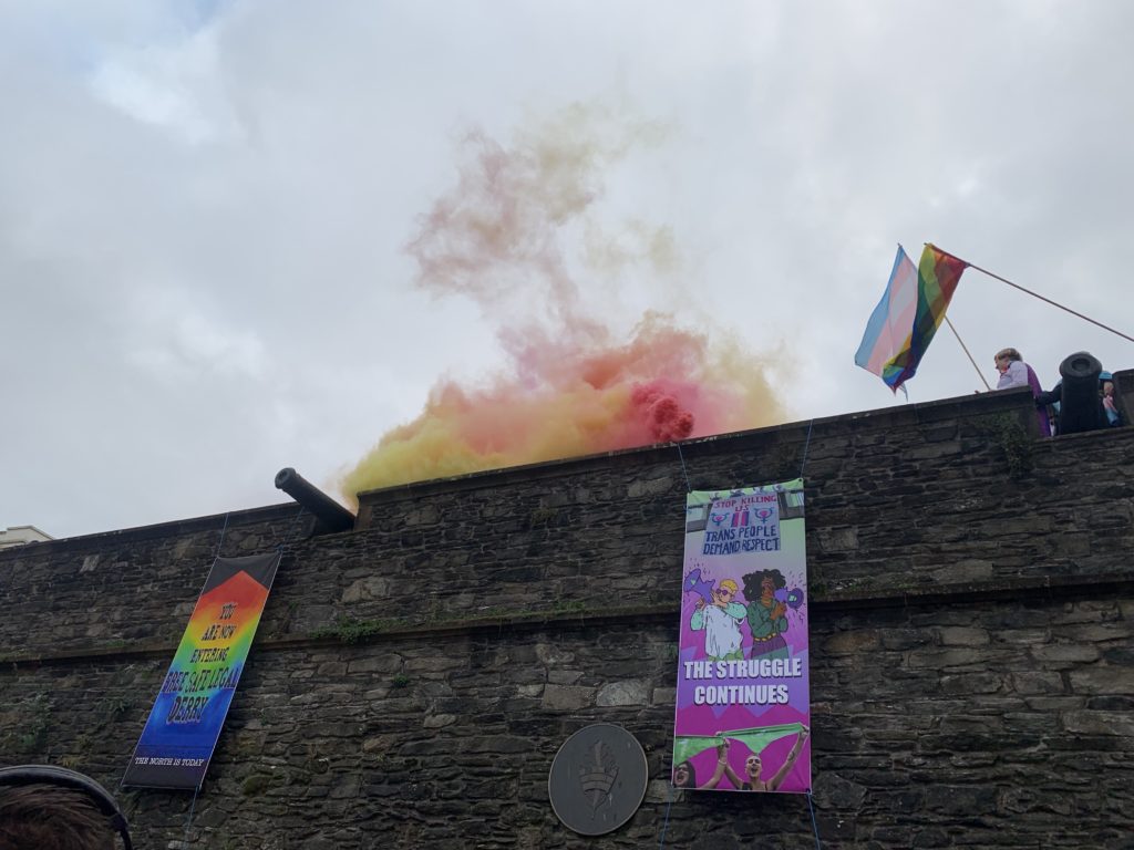 A view of the Derry walls from the Guildhall square. Festive pink and yellow smoke rises from the walls, and a rainbow and trans (blue, pink, and white) flag wave to the right of the image. Banners hang from the walls that say "You are now entering Free, Safe, Legal Derry" and "Trans People Demand Respect - The Struggle Continues"