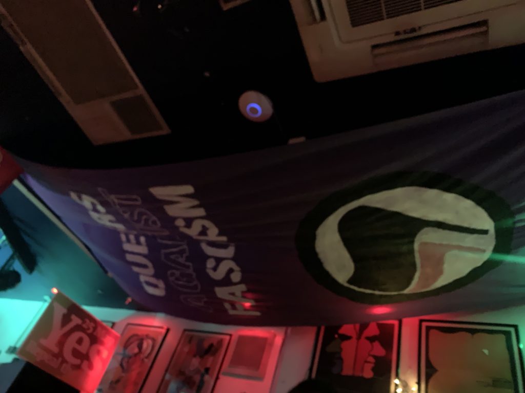 A dimly lit bar ceiling with a large banner draped overhead. It reads, "Queers Against Fascism" and shows the Antifa symbol - black and red flags against a white circular background. Underneath the banner is a "Yes" sign from the Together for Yes campaign in the Republic of Ireland.